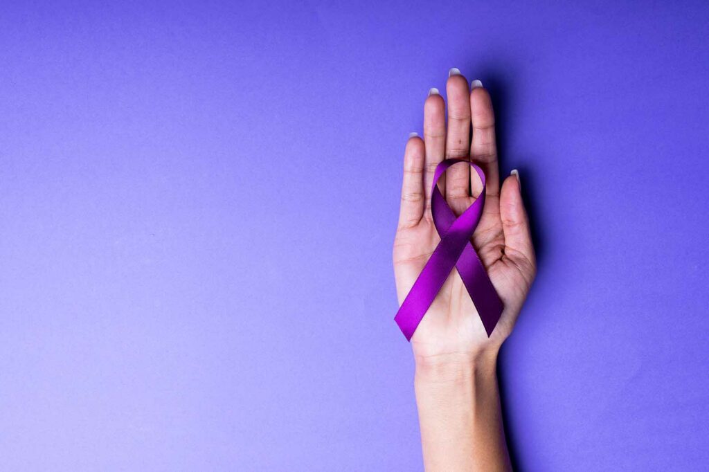 A woman's hand holding a purple ribbon representing pancreatic cancer on a purple background.